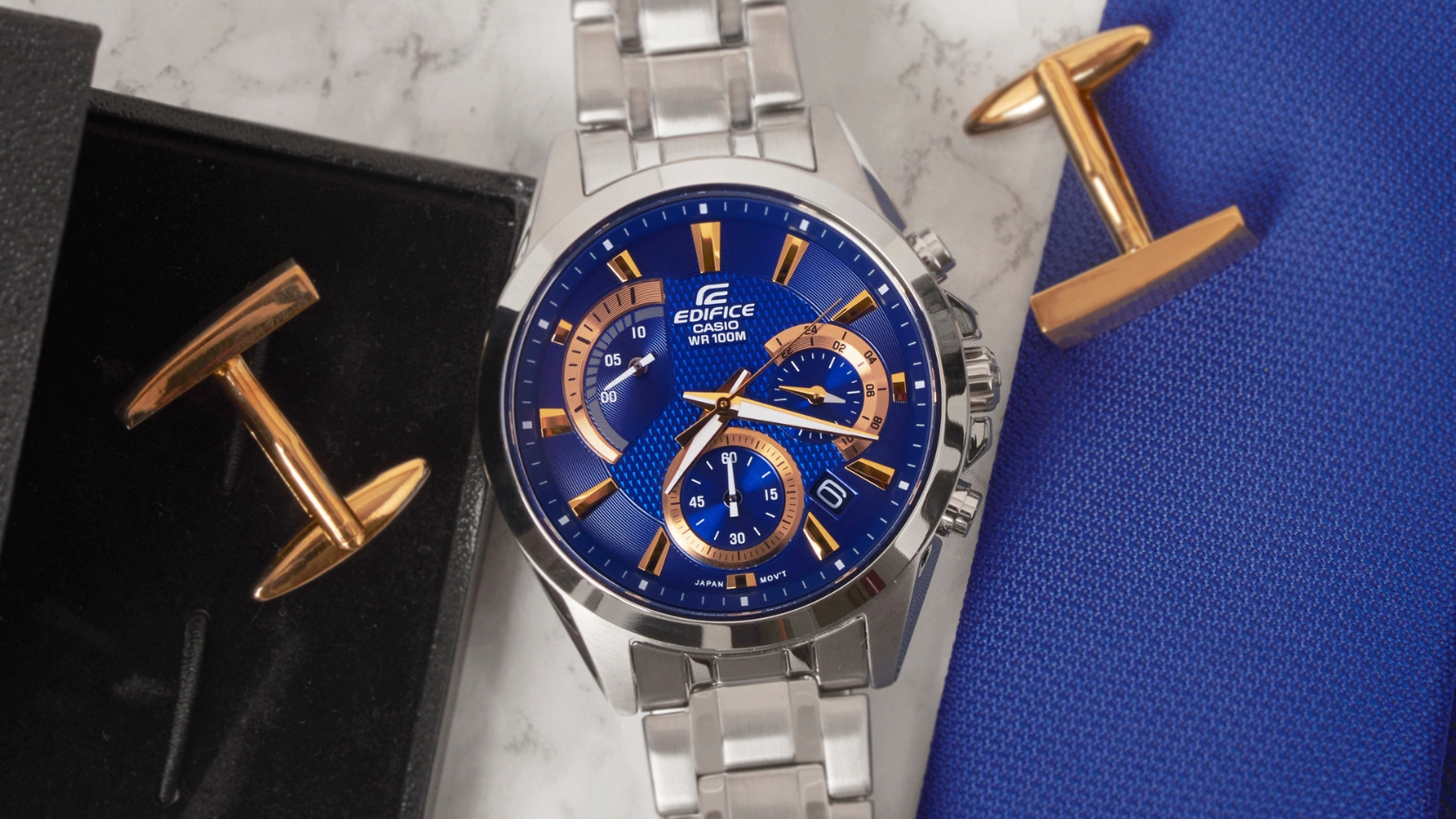 Casio Edifice z niebieską tarczą, złotymi wskazówkami i stalową bransoletą, obok dwóch spinek do mankietów na marmurze