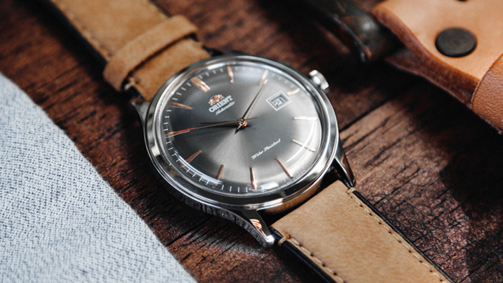 Orient Bambino z ciemnoszarą tarczą i datownikiem na jasnobrązowym skórzanym pasku, leżący na drewnianym blacie obok koszuli