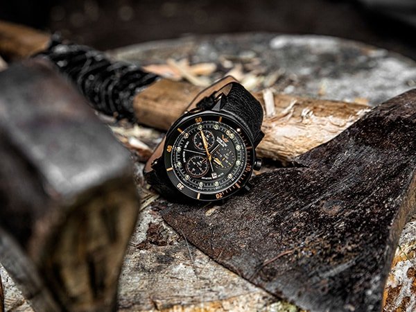 Męski zegarek na skórzanym czarnym pasku, leżący na drewnianym pniu obok starego siekierza.